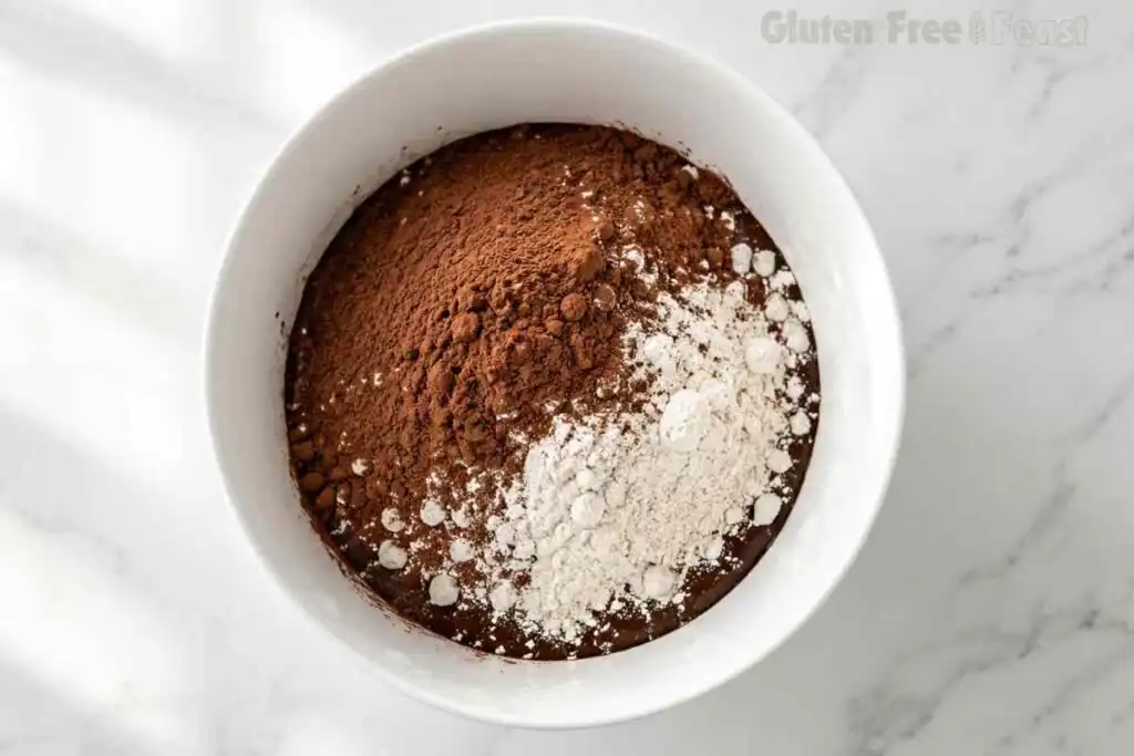 Gluten-free flour and cocoa powder added to brownie wet mixture in a white bowl