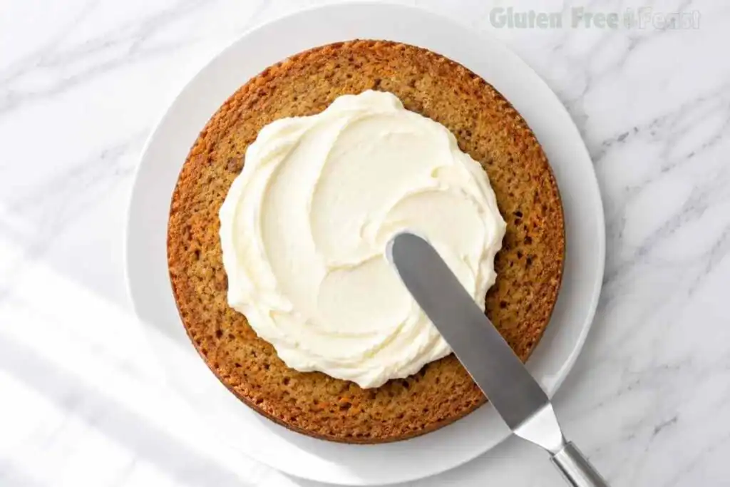 Cream cheese frosting being spread over a gluten-free carrot cake layer with an offset spatula.