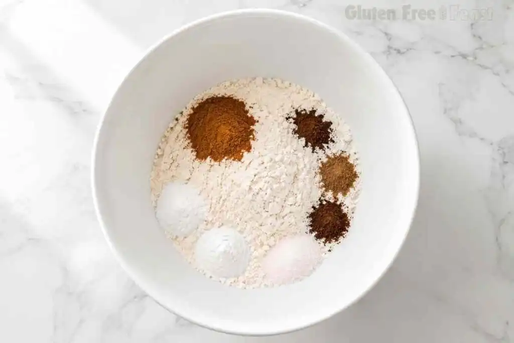 Gluten-free flour, cinnamon, nutmeg, cloves, allspice, baking soda, baking powder, and salt in a white bowl.