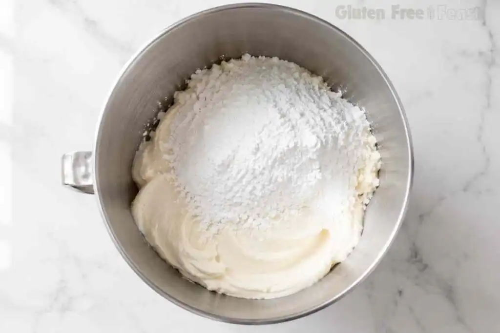 Powdered sugar added to cream cheese frosting in a stainless steel mixer bowl.