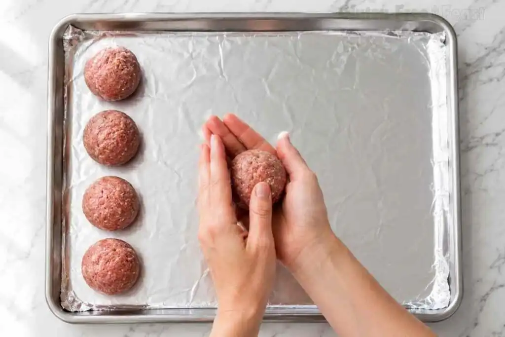 Hands rolling raw gluten free meatballs on a foil-lined baking sheet