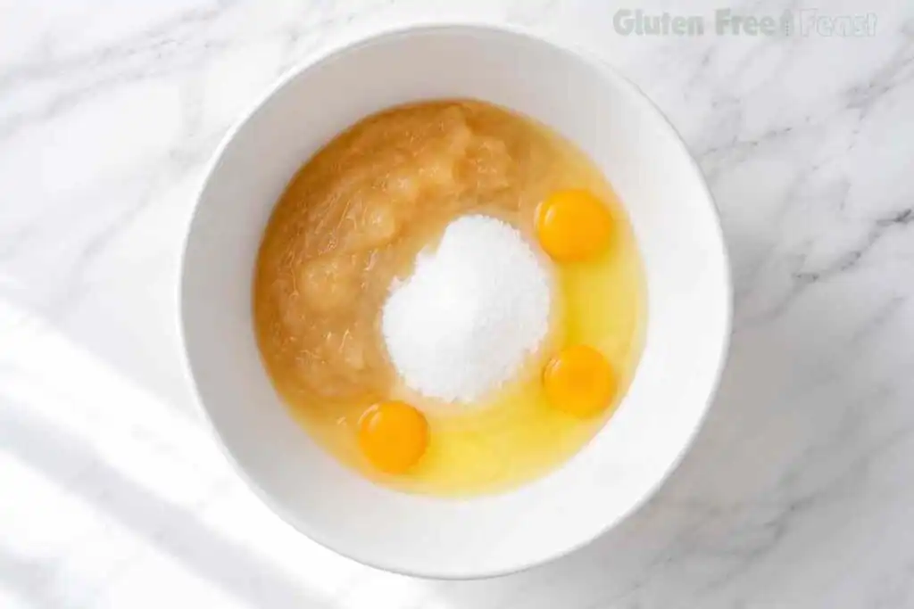 Eggs, applesauce, sugar, and vanilla in a white mixing bowl for gluten-free carrot cake.