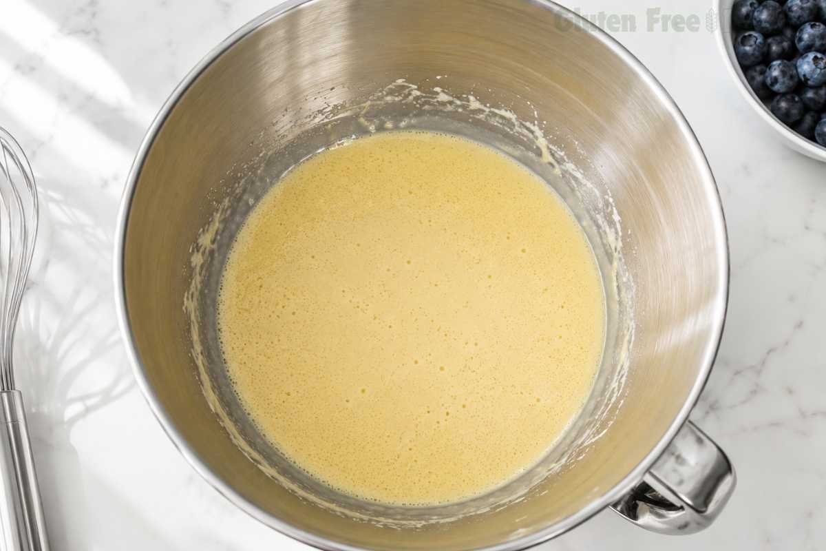 Overhead shot of eggs and sugar beaten to the ribbon stage — pale, airy and tripled in volume in a stainless steel stand mixer bowl on white marble — the structural base for gluten-free blueberry muffins