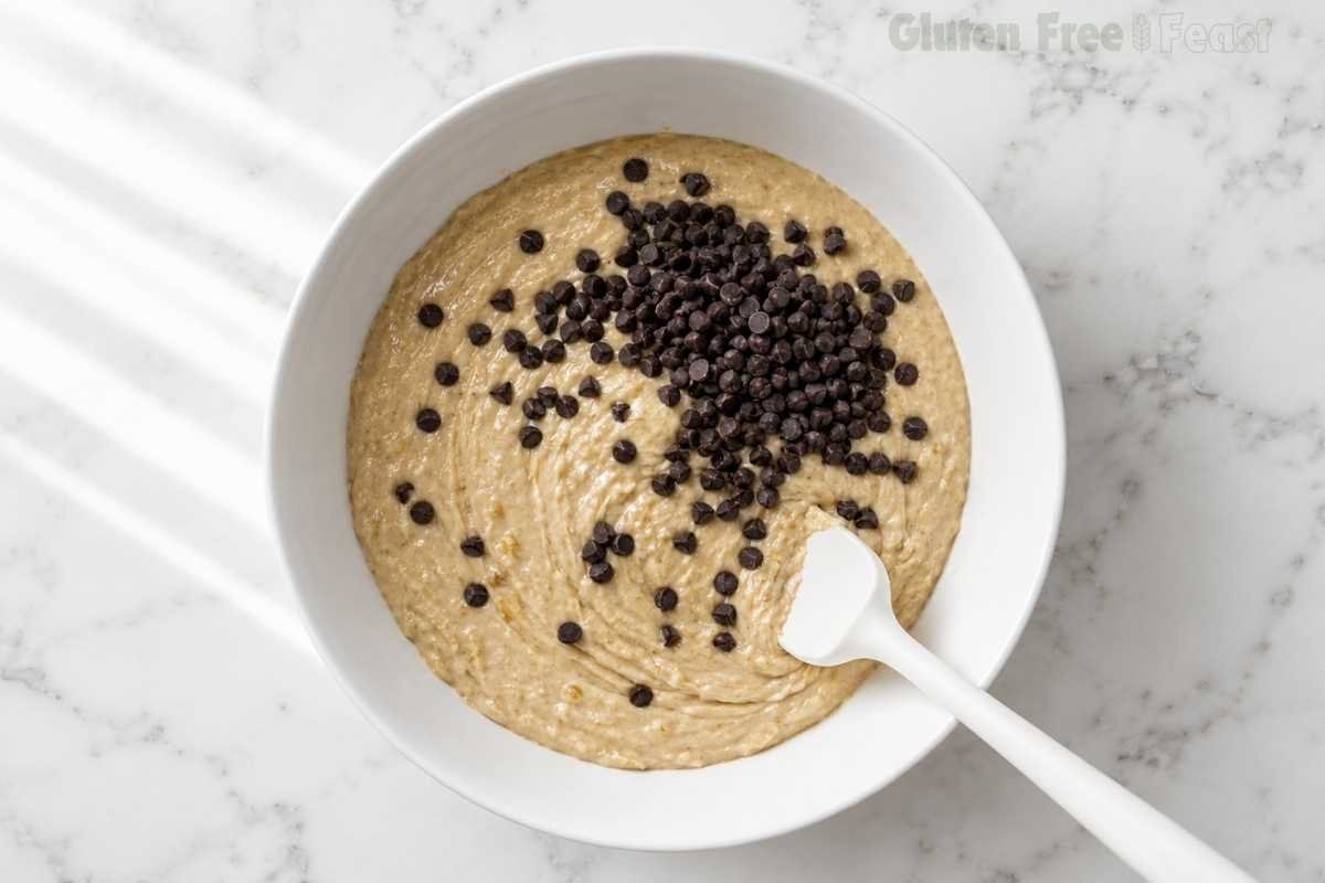 Overhead shot of gluten-free banana muffin batter in a white bowl with dark chocolate chips scattered on top being folded in with a white spatula on a marble countertop