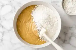White silicone spatula folding certified gluten free flour into banana bread batter in a white bowl on marble with extra flour bowl beside