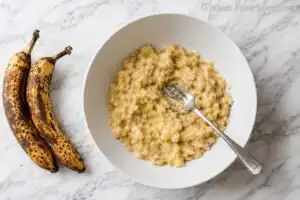 Two overripe spotted bananas beside a white bowl with mashed banana and a fork on a marble countertop for gluten free banana bread