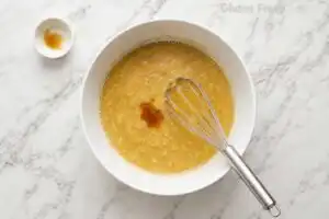 Golden banana bread wet mixture being whisked in a white ceramic bowl on marble with a small bowl of vanilla extract alongside