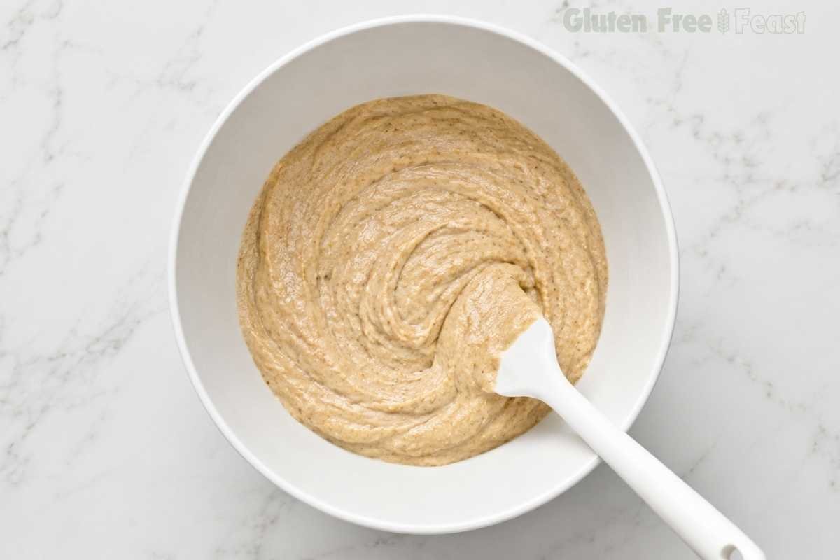 Overhead shot of thick finished gluten-free banana muffin batter in a large white bowl with a white spatula showing smooth scoopable consistency on white marble