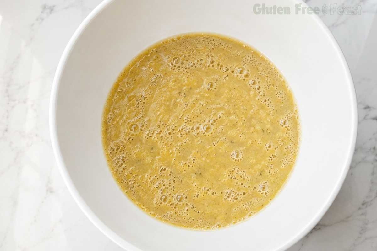 Overhead shot of smooth pale gluten-free banana muffin wet batter fully combined in a large white bowl on a marble countertop showing airy bubbly consistency
