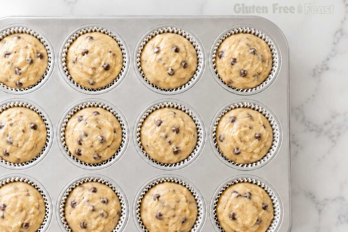 Close-up overhead shot of 12 gluten-free banana chocolate chip muffin cups fully filled and domed ready to go into the oven showing perfect portioning on white marble