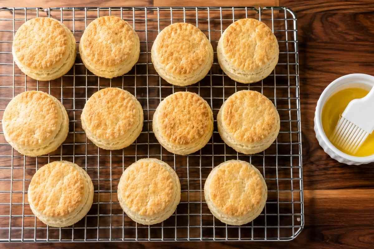 Baked gluten free buttermilk biscuits on a wire cooling rack next to a bowl of melted butter and a pastry brush