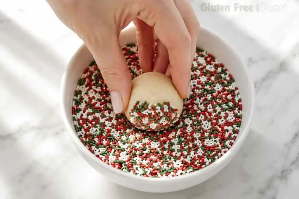 Dipping a gluten free Christmas cookie dough ball into a bowl of red green and white sprinkles