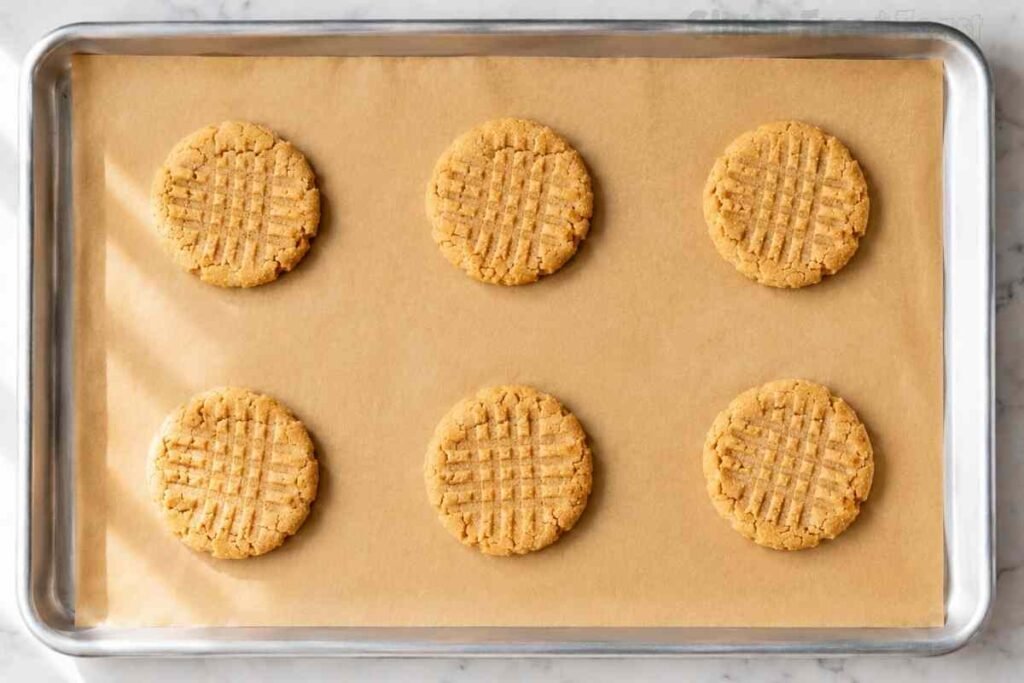 Six gluten free peanut butter cookies just baked for 10 to 12 minutes at 375F with lightly golden edges and fork cross-hatch pattern on parchment lined silver baking sheet cooling on marble