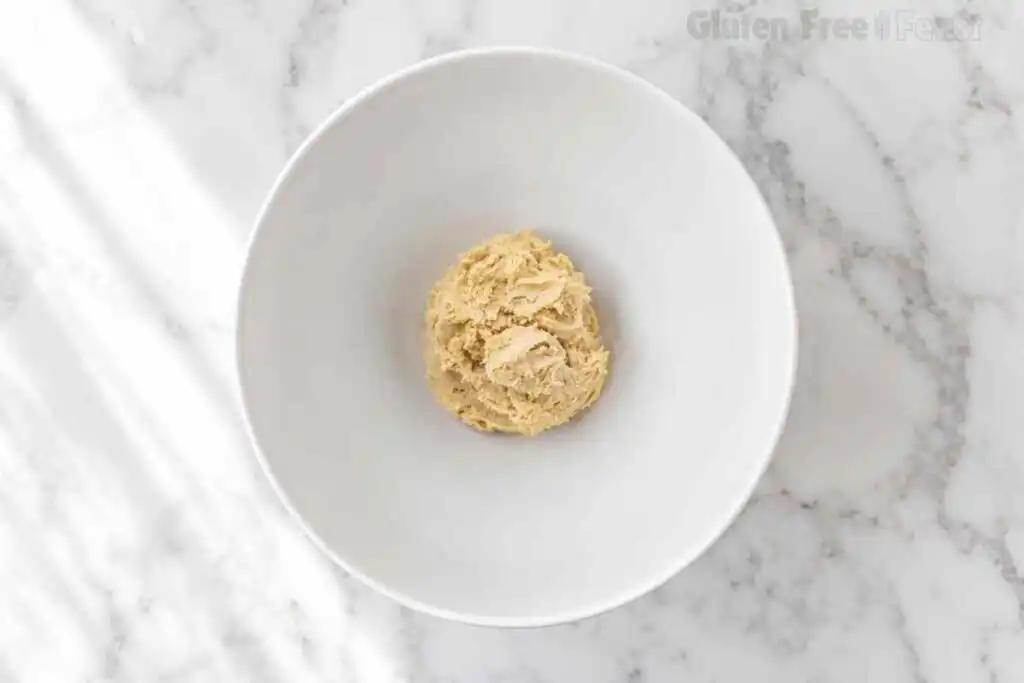 Finished gluten free sugar cookie dough ball in a white bowl, soft and pale golden after combining wet and dry ingredients