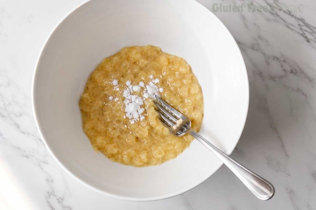 Overhead shot of mashed overripe bananas in a white ceramic bowl with a fork on a white marble surface, ready to be mixed into gluten-free banana muffin batter