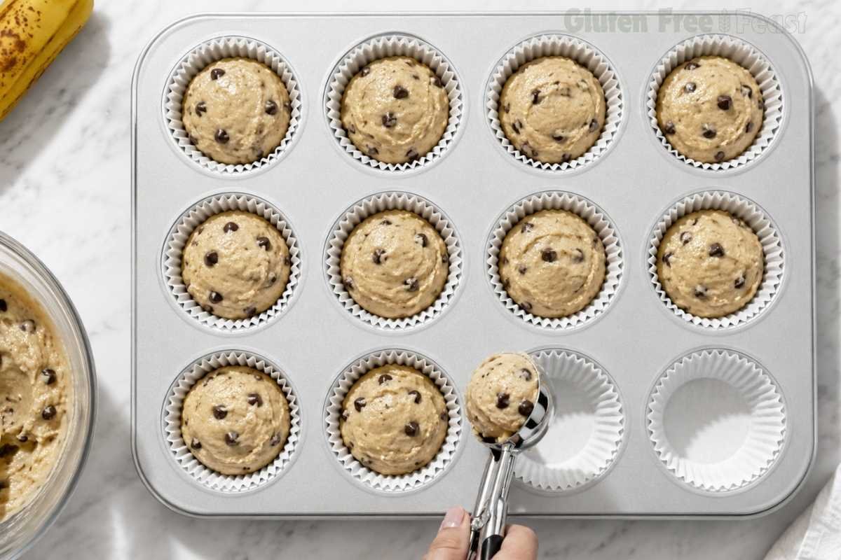 Overhead action shot of a cookie scoop filling the last cups of a muffin pan with gluten-free banana chocolate chip batter, showing uniform portioning technique on a marble countertop