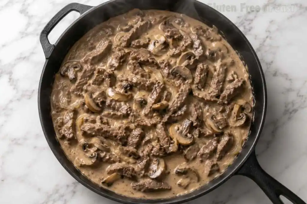 Creamy Gluten Free Beef Stroganoff cooking in a cast iron skillet with mushrooms and tender beef using GFCO-certified gluten-free ingredients.