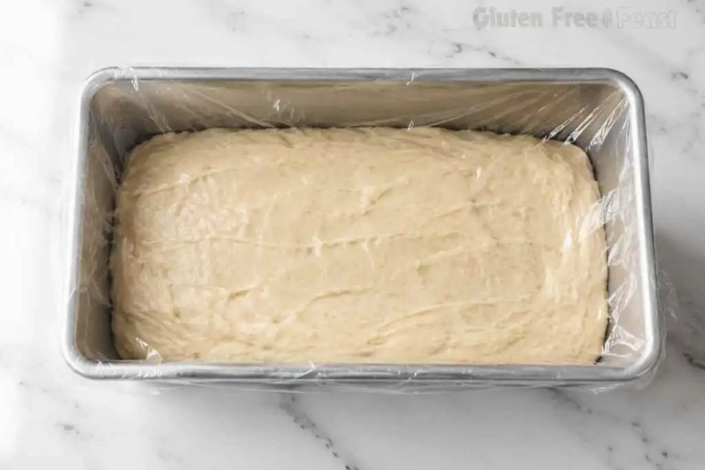 Gluten free bread batter spread inside a loaf pan and covered for proofing.