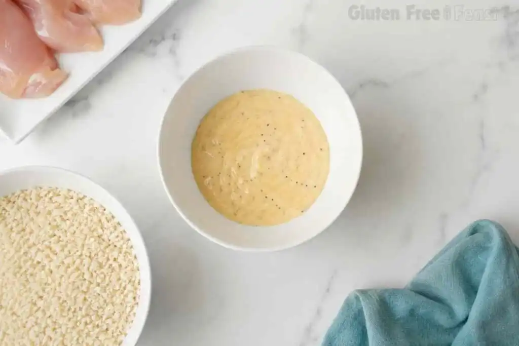 Creamy gluten-free chicken coating mixture in a white bowl with panko breadcrumbs and raw chicken nearby.