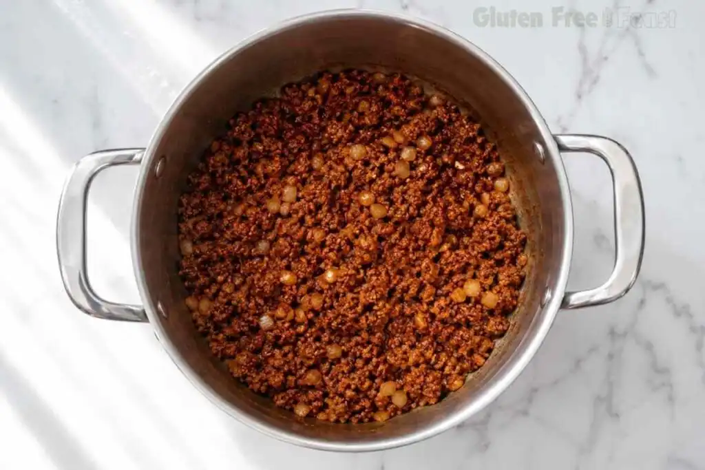 Ground beef mixed with chili spices and tomato paste in a stainless steel pot.